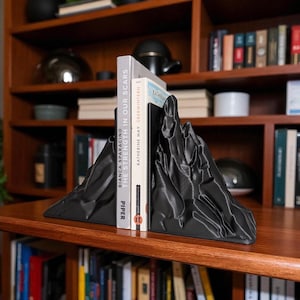 May include: Black mountain-shaped bookends holding two books on a wooden shelf. The bookends have a textured surface and are designed to resemble a mountain range. The books are titled "The Strength in Our Scars" and "Katherine May Oberwinter".