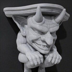 K&ouml;nnte beinhalten: Eine hellgraue, geschnitzte Regalhalterung mit einer d&auml;monischen Figur. Die Figur hat H&ouml;rner, ein breites Grinsen und hockt, um das Regal dar&uuml;ber zu st&uuml;tzen. Das Regal hat eine dekorative, geschwungene Kante. Die Halterung ist aus Holz.