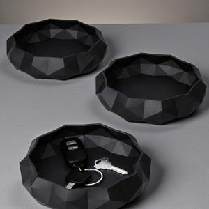 K&ouml;nnte beinhalten: Drei schwarze geometrische Tabletts mit facettiertem Design. Ein Tablett enth&auml;lt einen Satz Schl&uuml;ssel und einen Autoschl&uuml;sselanh&auml;nger. Die Tabletts bestehen aus mattem schwarzem Material und sind auf einer grauen Oberfl&auml;che angeordnet.