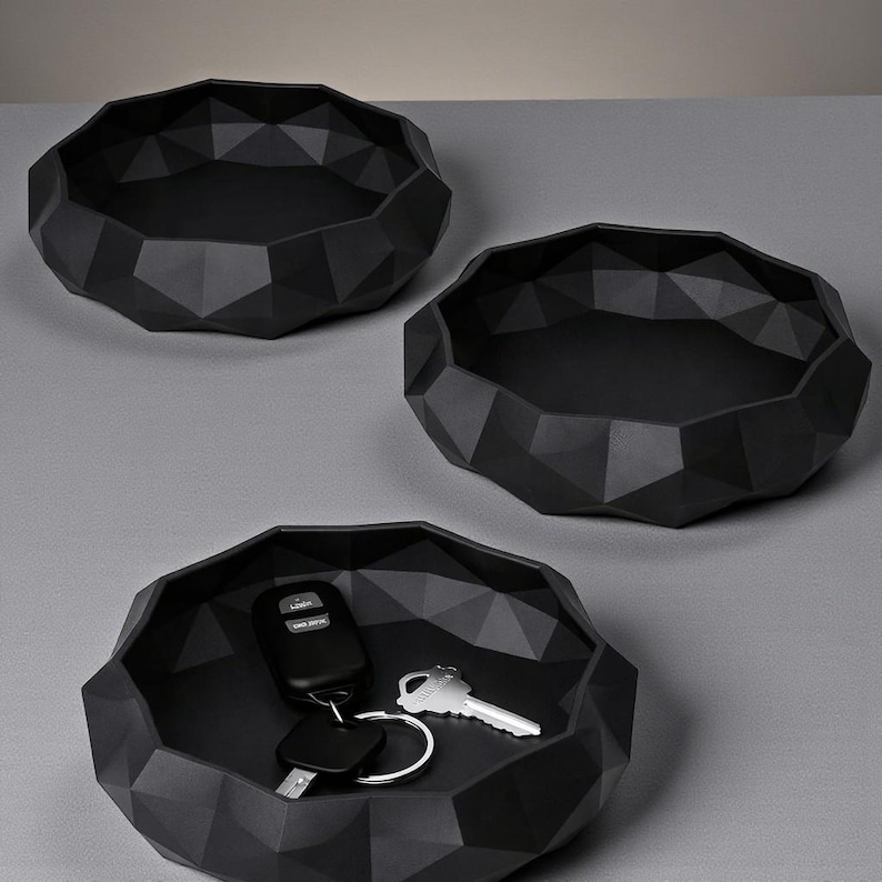 K&ouml;nnte beinhalten: Drei schwarze geometrische Tabletts mit facettiertem Design. Ein Tablett enth&auml;lt einen Satz Schl&uuml;ssel und einen Autoschl&uuml;sselanh&auml;nger. Die Tabletts bestehen aus mattem schwarzem Material und sind auf einer grauen Oberfl&auml;che angeordnet.