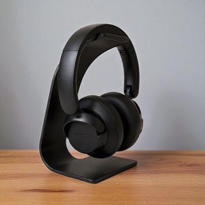 May include: Black over-ear headphones resting on a matching black headphone stand. The stand has a curved design and a flat base. The headphones are positioned to showcase their design and features. The background is a neutral gray.