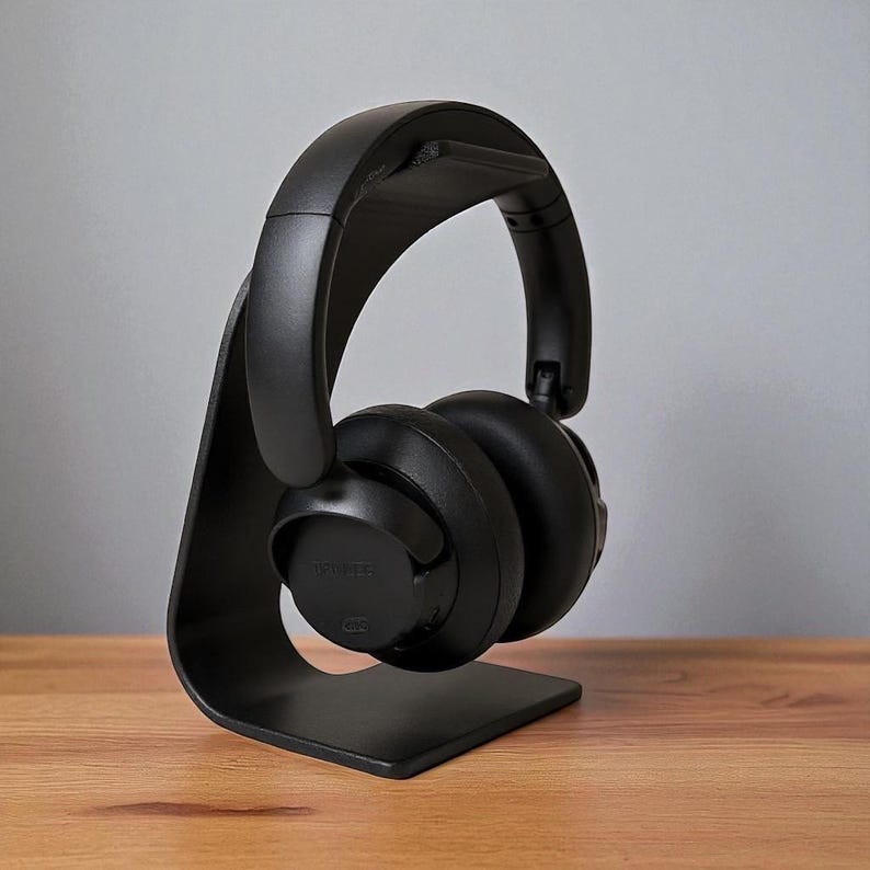 May include: Black over-ear headphones resting on a matching black headphone stand. The stand has a curved design and a flat base. The headphones are positioned to showcase their design and features. The background is a neutral gray.