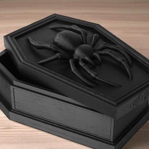 May include: Black coffin-shaped box with a raised spider design on the lid. The box is open, revealing the interior. The spider is detailed with eight legs and a segmented body. The box is made of wood and has a matte finish.