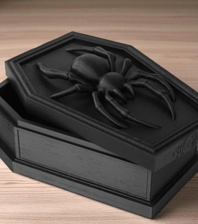May include: Black coffin-shaped box with a raised spider design on the lid. The box is open, revealing the interior. The spider is detailed with eight legs and a segmented body. The box is made of wood and has a matte finish.