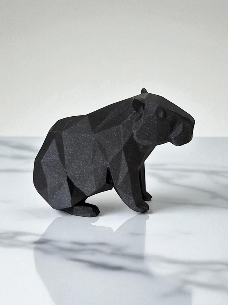 May include: A black, geometric capybara figurine. The sculpture is made of matte black material and features a faceted design, giving it a modern, angular appearance. The capybara is depicted in a seated position.