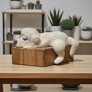 May include: A cream-colored dog sculpture resting on a wooden block. The dog has a relaxed pose with its head down and tail hanging off the side. The dog's nose is painted dark blue. The wooden block is rectangular and has a natural wood grain.