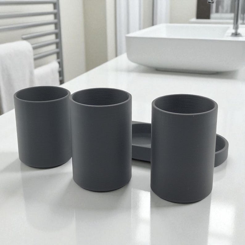 May include: Three grey cylindrical bathroom tumblers and a small tray sit on a white countertop. The tumblers are evenly spaced and have a smooth, matte finish. The tray is rectangular with rounded edges.