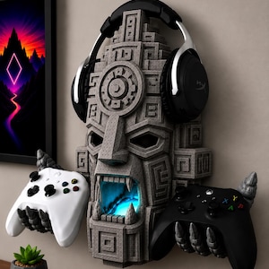 May include: A gray stone-textured wall mount shaped like a carved mask with an open mouth glowing with blue light. The mask features two stone hands extending outward to hold a white game controller and a black game controller, with black headphones resting on top.