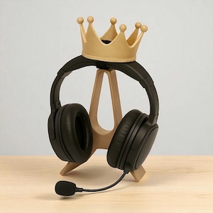 May include: Black over-ear headphones with a microphone, resting on a wooden stand. A gold-colored crown sits atop the headphones. The stand is made of light-colored wood and is on a wooden surface.