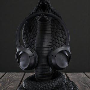 Op de afbeelding: Een zwarte koptelefoonstandaard in de vorm van een cobra met een zwarte JBL koptelefoon. De standaard heeft een getextureerde afwerking en de koptelefoon is geplaatst alsof hij door de cobra wordt gedragen. De achtergrond is donker. De standaard is gemaakt van een getextureerd materiaal.