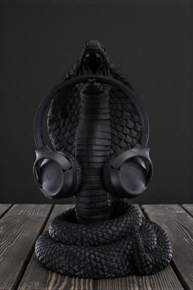 Op de afbeelding: Een zwarte koptelefoonstandaard in de vorm van een cobra met een zwarte JBL koptelefoon. De standaard heeft een getextureerde afwerking en de koptelefoon is geplaatst alsof hij door de cobra wordt gedragen. De achtergrond is donker. De standaard is gemaakt van een getextureerd materiaal.