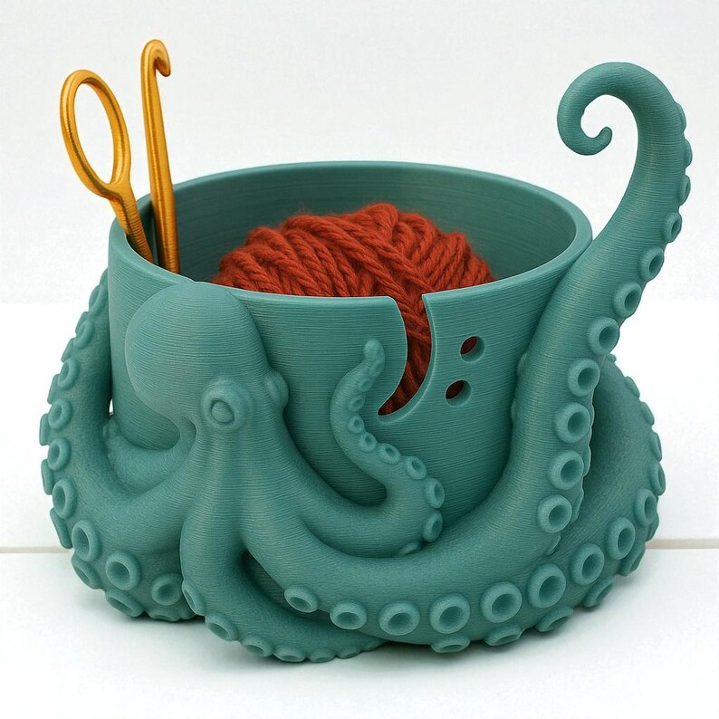 May include: A teal octopus-shaped yarn bowl with a ball of orange yarn inside. Two gold crochet hooks are placed in the bowl. The bowl has a unique design with tentacles and a curved tentacle extending upwards.