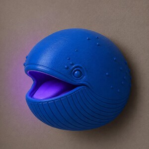 Op de afbeelding: Een blauwe walvisvormige wandlamp met een open mond en een paars interieur. De walvis heeft een getextureerd oppervlak met kleine bobbels en verticale lijnen. De lamp is gemonteerd op een neutraal gekleurde muur.