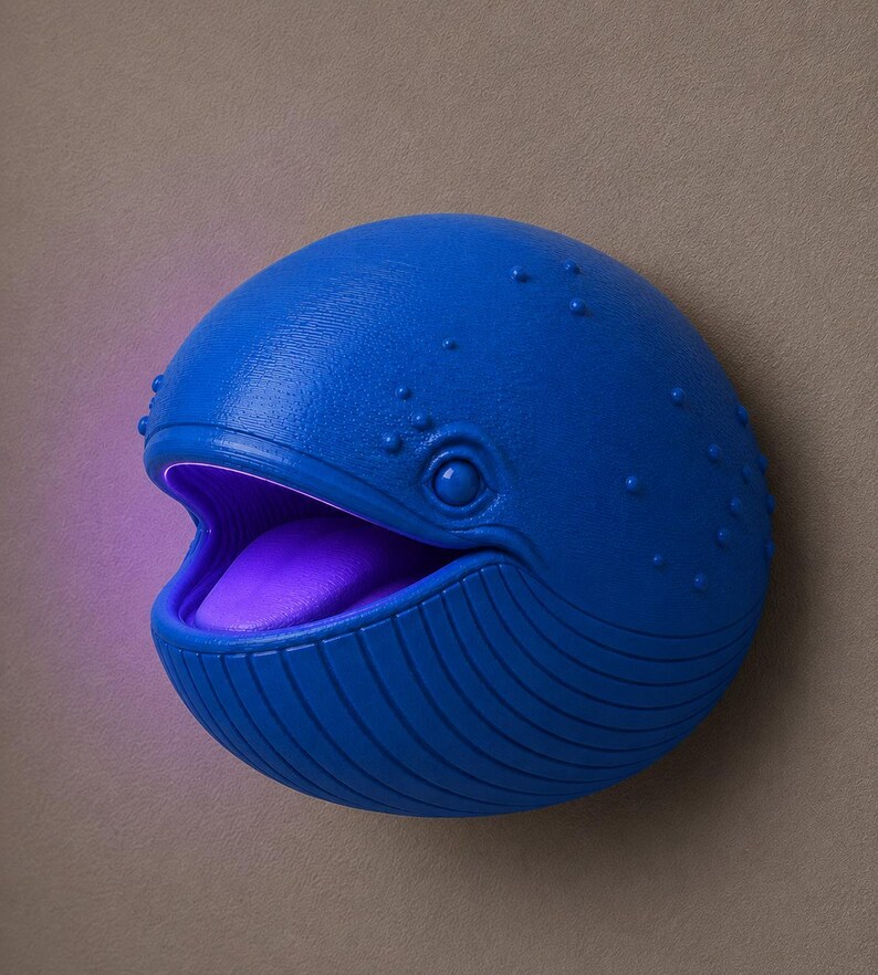 Op de afbeelding: Een blauwe walvisvormige wandlamp met een open mond en een paars interieur. De walvis heeft een getextureerd oppervlak met kleine bobbels en verticale lijnen. De lamp is gemonteerd op een neutraal gekleurde muur.