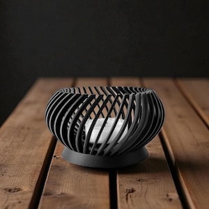 Pu&ograve; includere: Un portacandele nero, sferico, con un design a spirale. Il supporto poggia su una base rotonda e contiene una candela bianca. Il portacandele &egrave; su una superficie di legno.