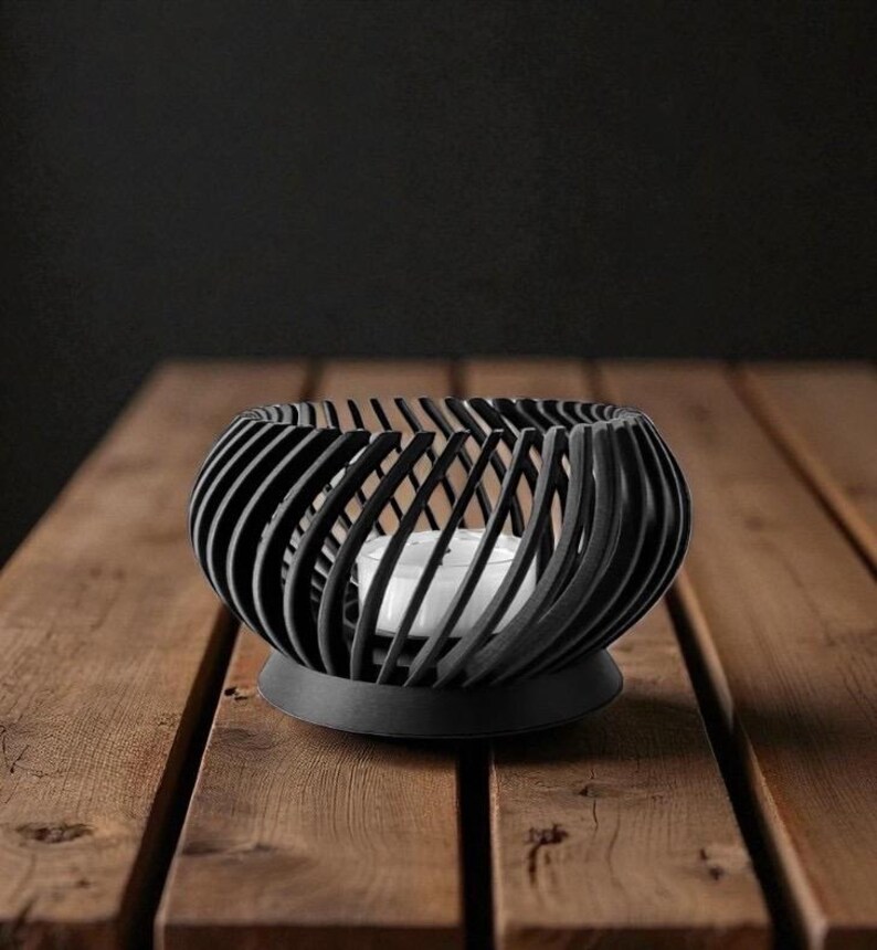 Pu&ograve; includere: Un portacandele nero, sferico, con un design a spirale. Il supporto poggia su una base rotonda e contiene una candela bianca. Il portacandele &egrave; su una superficie di legno.