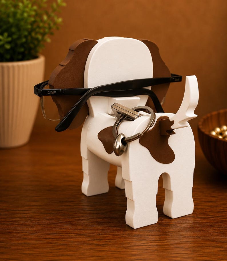 May include: A wooden dog-shaped eyeglasses and key holder. The holder is white with brown spots and ears. Black eyeglasses rest on the dog's head, and a set of keys hangs from the tail. The holder is on a wooden surface.