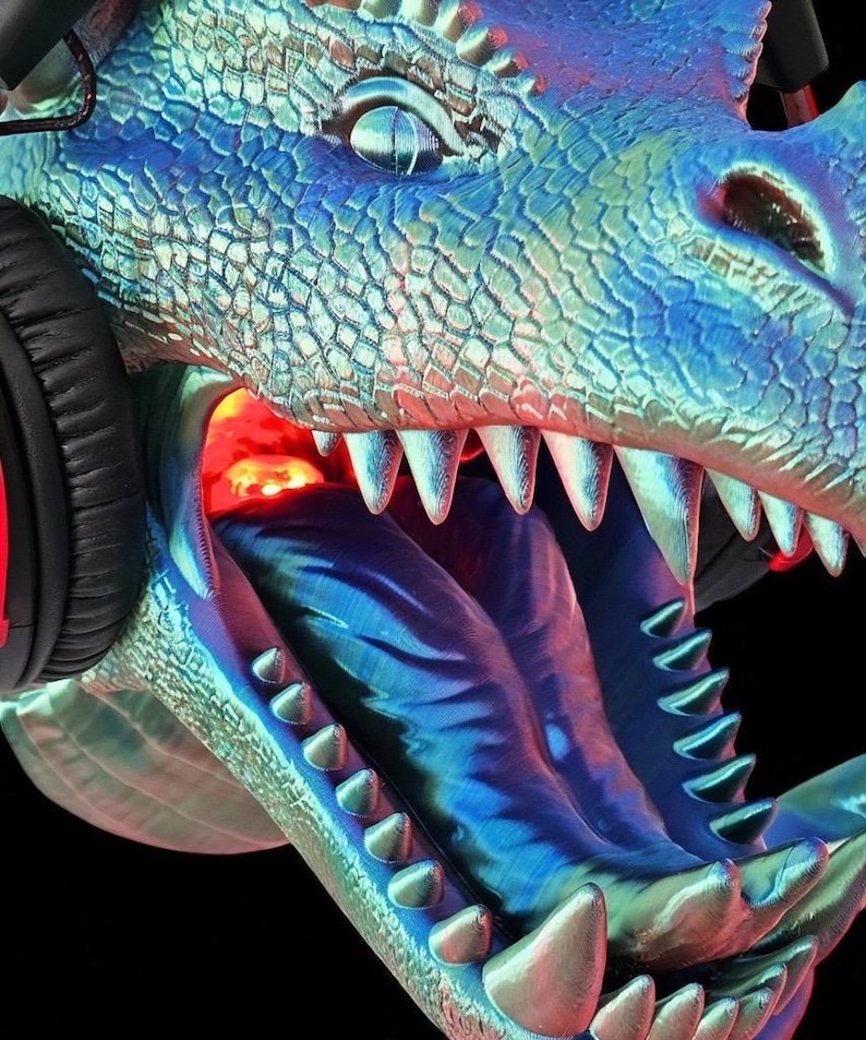 May include: A detailed, close-up image of a dragon's head with a headset. The dragon is rendered in shades of blue and green with sharp teeth and an open mouth. The headset is black with red accents. The background is black.