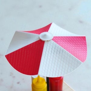 May include: A miniature red, white, and pink striped umbrella, likely for a food display. The umbrella is mounted on a wooden base with yellow, red, and black condiment bottles. The umbrella has a white button in the center.