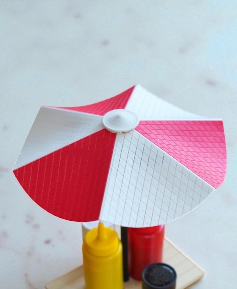 May include: A miniature red, white, and pink striped umbrella, likely for a food display. The umbrella is mounted on a wooden base with yellow, red, and black condiment bottles. The umbrella has a white button in the center.