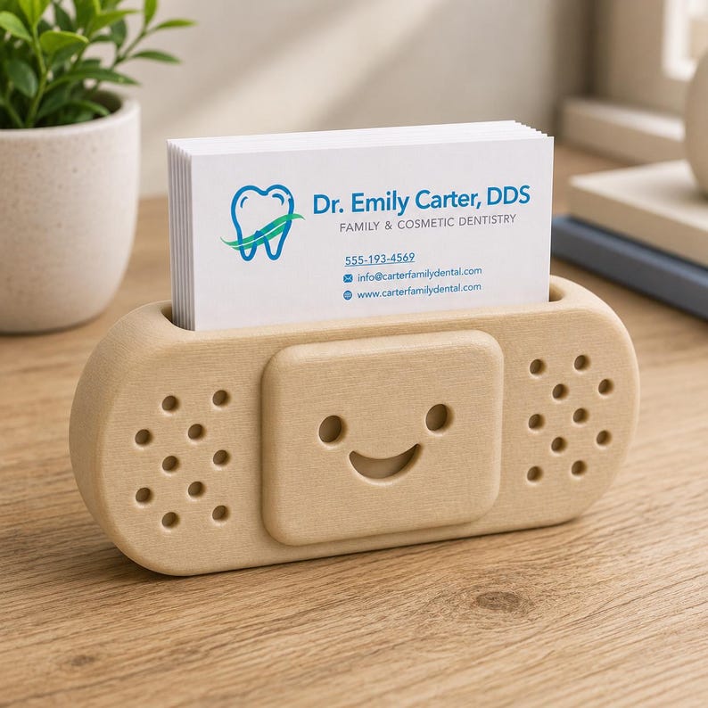 Op de afbeelding: Een lichtgekleurde houten visitekaarthouder in de vorm van een pleister, met een lachend gezicht. De houder bevat een stapel witte visitekaartjes met de tekst "Dr. Emily Carter, DDS, Family & Cosmetic Dentistry."