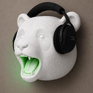 May include: A white panda head sculpture with black headphones. The panda's mouth is open, revealing a glowing green interior. The headphones are black with the brand name "HyperX" visible on the ear cup.