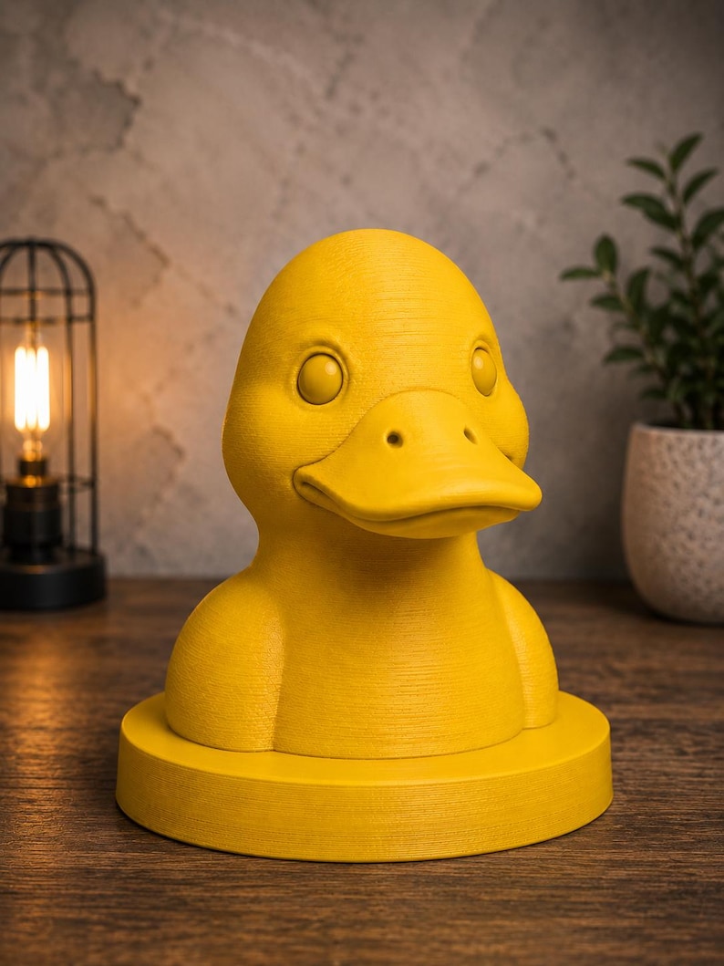 May include: A bright yellow duck-shaped decorative object. The duck has a detailed textured surface, with visible eyes and a beak. It is mounted on a circular base. The background includes a lamp and a potted plant, suggesting a home decor theme.