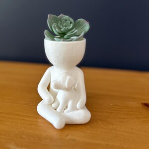 Puede incluir: Una figura de cer&aacute;mica blanca de una persona sentada con un perro, sosteniendo una planta suculenta en lugar de una cabeza. La planta tiene hojas verdes y grises. La figura est&aacute; sobre una superficie de madera.