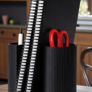 May include: A black desk tidy with three sections. It contains spiral-bound notebooks, a pen, and red scissors. The organiser is made of a ridged material and is on a wooden surface. The background shows a kitchen.