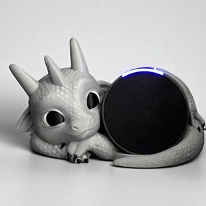 May include: A gray dragon-shaped speaker holder with a round black speaker. The dragon has large eyes, horns, and a textured body. The speaker has a blue light on top. The dragon is lying down.