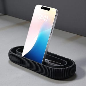 May include: A black phone stand with a ribbed design, holding a silver smartphone displaying a colorful screen. A stylus rests in the stand's inner compartment. The time 9:41 is visible on the phone's screen. The stand is on a light gray surface.