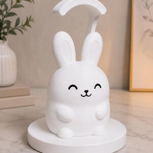May include: A white bunny-shaped night light with a curved top, smiling face, and a round base. The lamp is designed to sit on a table or desk, providing soft illumination. The lamp is made of a smooth, matte material.