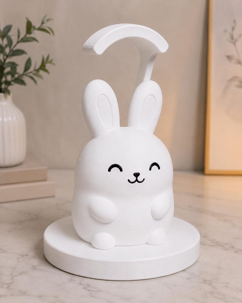 May include: A white bunny-shaped night light with a curved top, smiling face, and a round base. The lamp is designed to sit on a table or desk, providing soft illumination. The lamp is made of a smooth, matte material.
