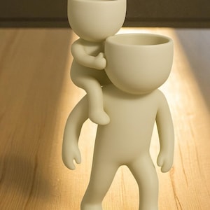 Op de afbeelding: Een cr&egrave;mekleurige keramische plantenbak in de vorm van een persoon die een andere persoon op zijn schouders draagt. De bovenkant van elke figuur is een komvormige plantenbak. De figuren hebben eenvoudige, afgeronde kenmerken en staan op een houten oppervlak.