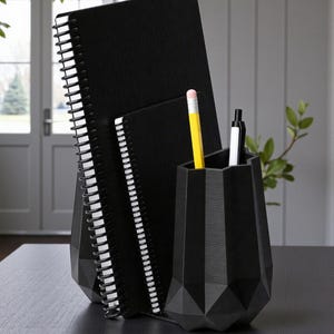 May include: A black desk organizer set featuring two spiral-bound notebooks and a geometric pen holder. A yellow pencil and two pens are in the holder. The notebooks are standing upright. The set is on a dark surface.