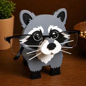 May include: A gray and white raccoon-shaped glasses holder. The wooden holder has black ears, eyes, and nose, with white accents. The raccoon is wearing a pair of black-framed glasses. The holder is sitting on a wooden surface.