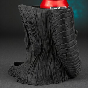 May include: A black, textured, 3D-printed can holder shaped like a tree trunk with a snake-like arm. A red can with white text is inside the holder. The holder is on a dark background.
