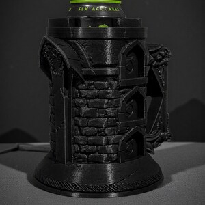 May include: Black 3D-printed beverage can holder with a castle tower design. The holder features a brick pattern, arched windows, and decorative elements. The top of the can is visible, with the text "SEM A&Ccedil;UCARES" in green.
