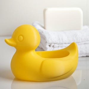 Op de afbeelding: Een felgele zeepbakje in de vorm van een eend. De eend heeft een rond lichaam, een kleine snavel en een cirkelvormige opening om zeep in te doen. Witte handdoeken en een stuk zeep staan op de achtergrond.