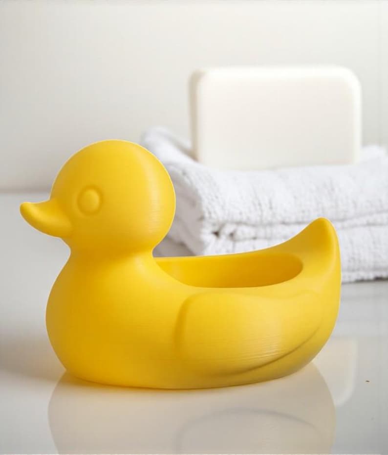 Op de afbeelding: Een felgele zeepbakje in de vorm van een eend. De eend heeft een rond lichaam, een kleine snavel en een cirkelvormige opening om zeep in te doen. Witte handdoeken en een stuk zeep staan op de achtergrond.