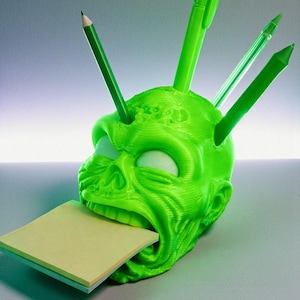 May include: A neon green, 3D-printed skull-shaped desk organizer. The skull holds a stack of sticky notes in its mouth and has several green pens and pencils protruding from the top. The design features a textured surface.