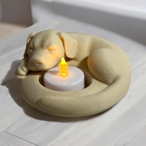 Puede incluir: Un portavelas con forma de perro de color crema con un dise&ntilde;o de Labrador durmiendo. El portavelas tiene una base circular con una vela blanca y una luz LED amarilla parpadeante. El perro est&aacute; acurrucado, creando un art&iacute;culo acogedor y decorativo.