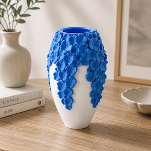 May include: A white ceramic vase with a blue floral design. The vase has a rounded shape with a narrow base and a wide opening. The blue flowers cascade down the sides of the vase, creating a textured appearance. The vase is a decorative home decor item.