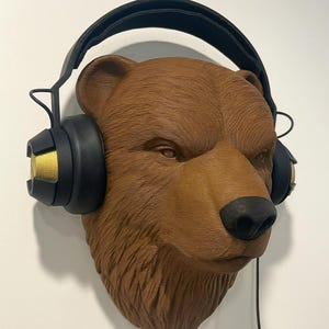 May include: A brown bear head sculpture with black headphones. The bear's fur is textured, and the headphones have gold accents. A black wire extends from the headphones. The sculpture is mounted on a white wall.