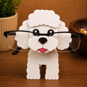 May include: A white poodle-shaped eyeglass holder with black rimmed glasses. The holder has a pink tongue and black eyes and nose. The eyeglass holder is made of a solid material and sits on a wooden surface.