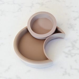 Puede incluir: Un juego de bandejas decorativas modernas y minimalistas en tonos beige y topo. El conjunto incluye un cuenco circular sobre una bandeja en forma de media luna y una bandeja semicircular m&aacute;s grande.