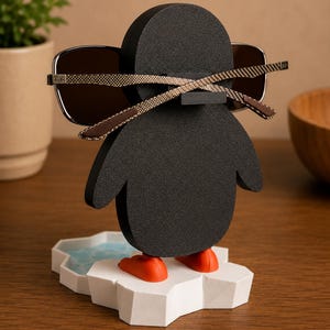 May include: A black penguin-shaped eyeglass holder with orange feet and a white and blue ice floe base. The holder is holding a pair of sunglasses with brown frames. The eyeglass holder is on a wooden surface.