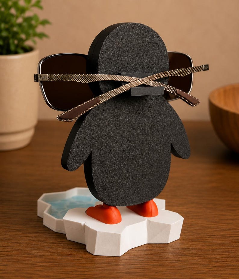 May include: A black penguin-shaped eyeglass holder with orange feet and a white and blue ice floe base. The holder is holding a pair of sunglasses with brown frames. The eyeglass holder is on a wooden surface.