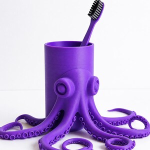 May include: A purple octopus-shaped toothbrush holder with a matching toothbrush. The holder has a cylindrical body and the octopus design wraps around it. The toothbrush has a black bristle head. The tentacles of the octopus are arranged around the base.