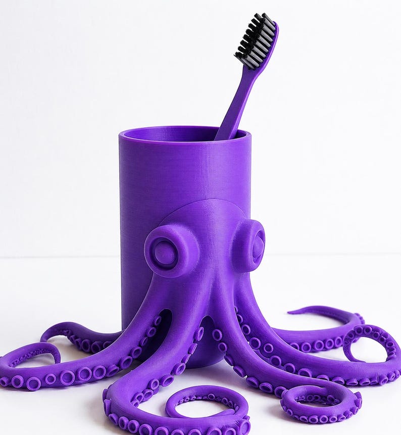 May include: A purple octopus-shaped toothbrush holder with a matching toothbrush. The holder has a cylindrical body and the octopus design wraps around it. The toothbrush has a black bristle head. The tentacles of the octopus are arranged around the base.